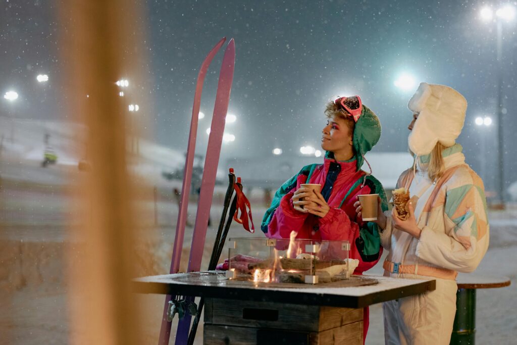 To unge i farverigt skitøj nyder varme drikke og mad ved bålfad i sneklædt vinterlandskab – DJ Gram tilbyder mobil DJ-service til skiture, vinterevents og afterski-fester i hele Danmark.