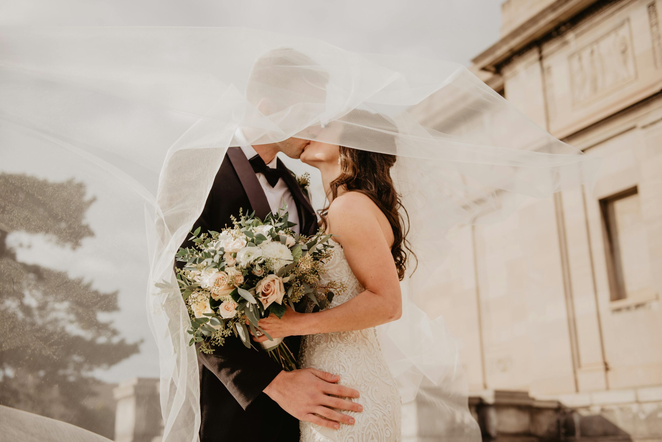 Brudepar kysser under sløret foran en historisk bygning med blomsterbuket i hånden – DJ Gram tilbyder professionel DJ-service til bryllupper og romantiske private events i hele Danmark.
