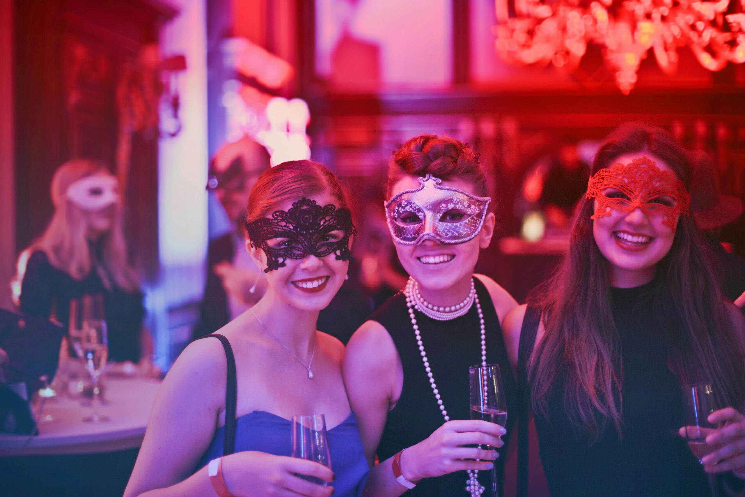 Tre unge kvinder med masker og festtøj smiler og holder champagneglas til en elegant maskeradefest – DJ Gram tilbyder professionel DJ-service til temafester, privatfester og eksklusive events i hele Danmark.