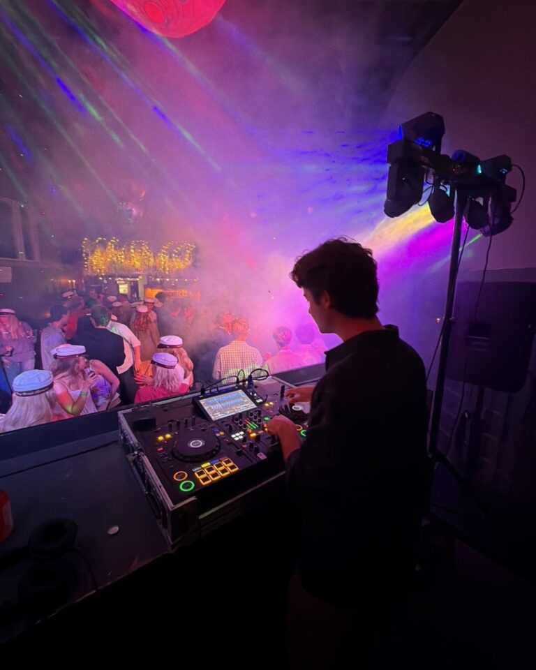 DJ at mixing console controlling event production with pink and purple laser beams, fog effects, and dancing crowd wearing hats inside tent venue