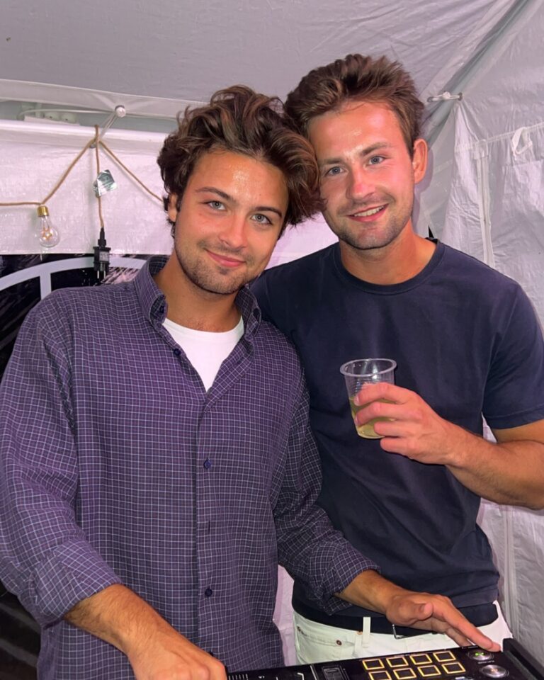 Two DJs posing together inside tent event space with mixing equipment visible, both smiling and enjoying beverages during entertainment performance