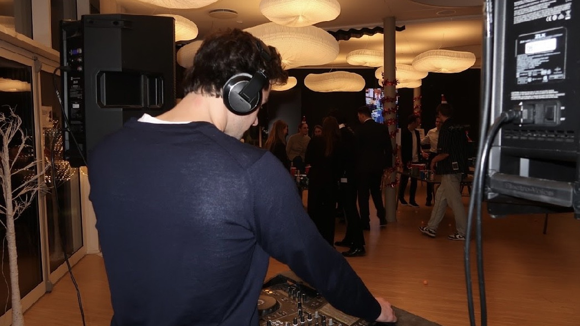 DJ with headphones at mixing console in modern indoor event space with warm ceiling lighting, guests socializing on wooden floor, contemporary décor