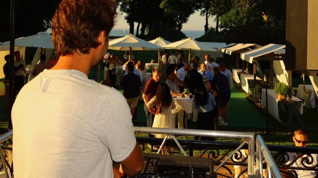 DJ overlooking luxury outdoor daytime event from elevated booth with white umbrellas, guest pavilions, manicured lawn, waterfront views, and guests mingling under clear sky
