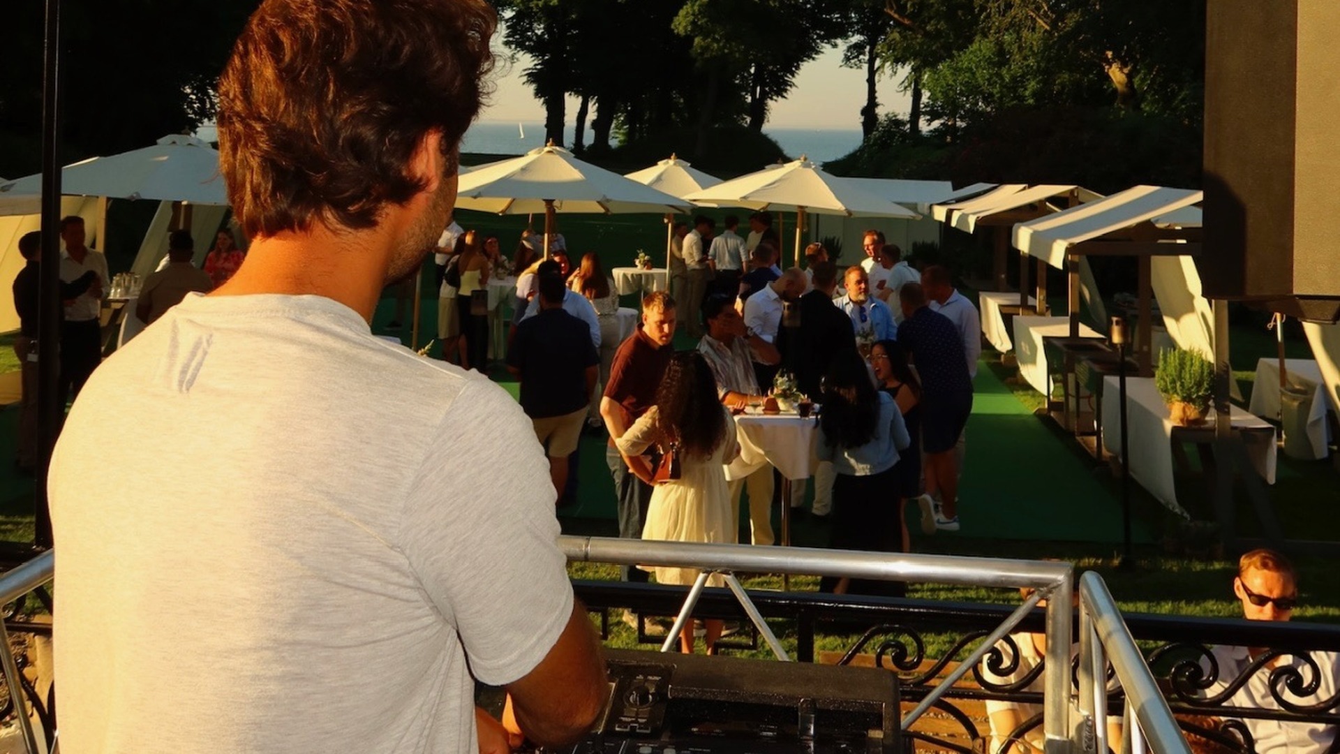 DJ overlooking luxury outdoor daytime event from elevated booth with white umbrellas, guest pavilions, manicured lawn, waterfront views, and guests mingling under clear sky