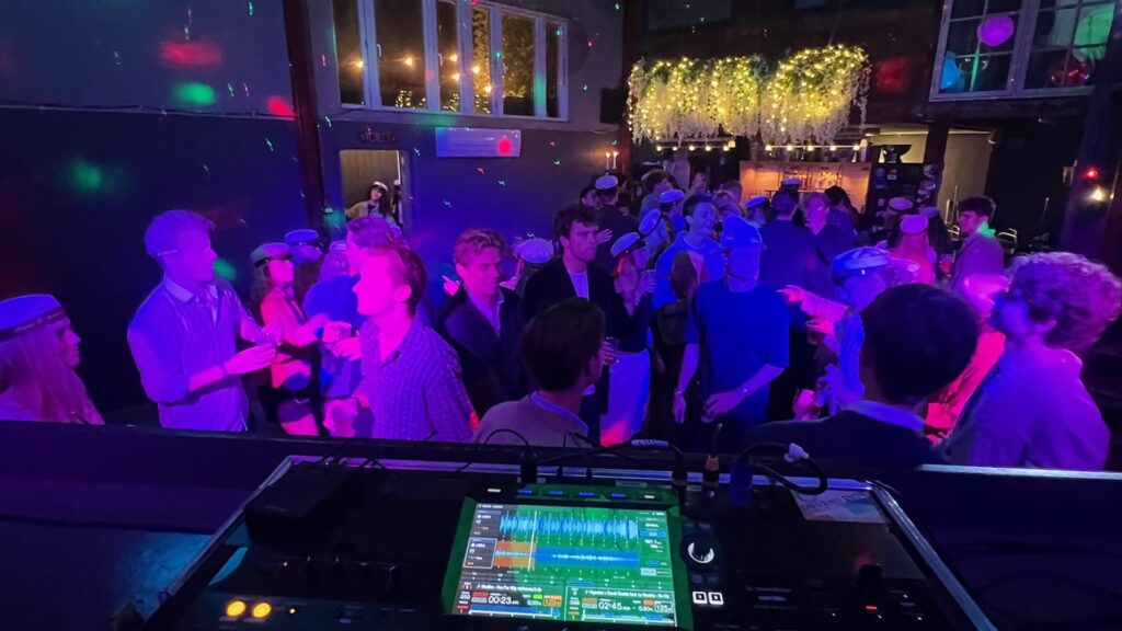 DJ mixer console at rooftop venue with large dancing crowd, dramatic magenta and blue lighting, gold glitter backdrop, and urban nighttime views from windows