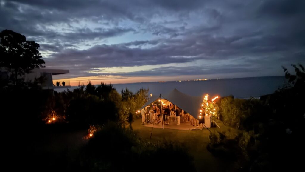 Illuminated marquee tent at dusk on waterfront with ambient lighting, guests gathering, dramatic twilight sky, ocean backdrop, and landscaped grounds