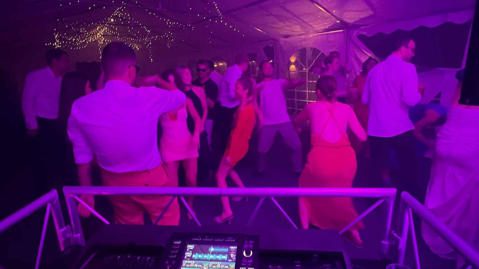 DJ mixing console at tent event with large dancing crowd, vibrant magenta and purple ambient lighting, decorative gold string lights overhead, packed dance floor