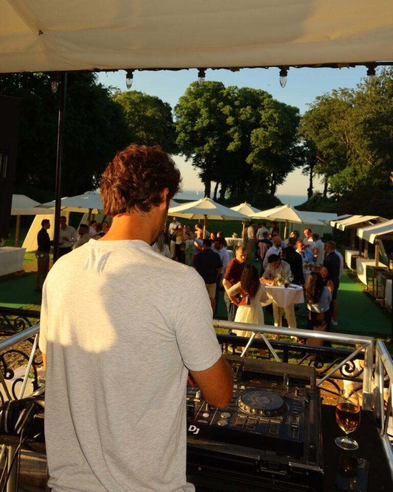 DJ at mixer console from elevated booth overlooking luxury outdoor event with white umbrellas, guests mingling, manicured lawn, tree-lined waterfront, and golden hour sunlight