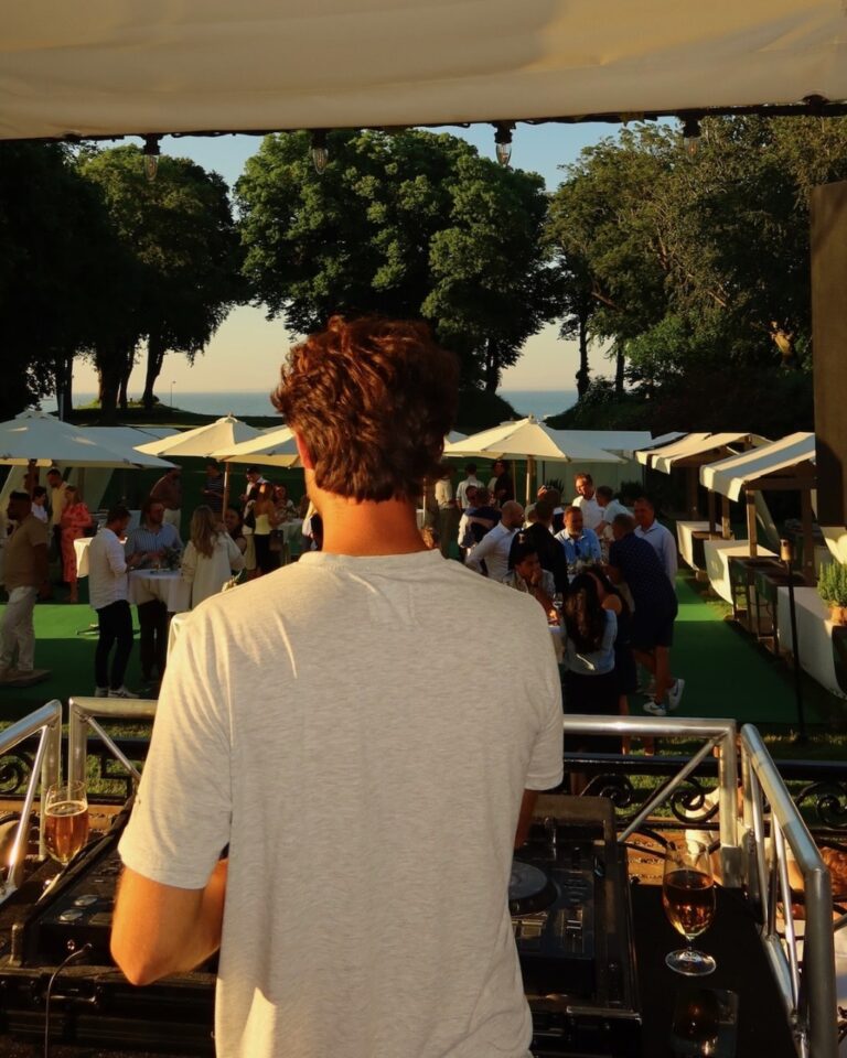 DJ overlooking luxury outdoor daytime event from elevated DJ booth with white umbrellas, guest pavilions, manicured lawn, trees, and waterfront views under bright sunshine