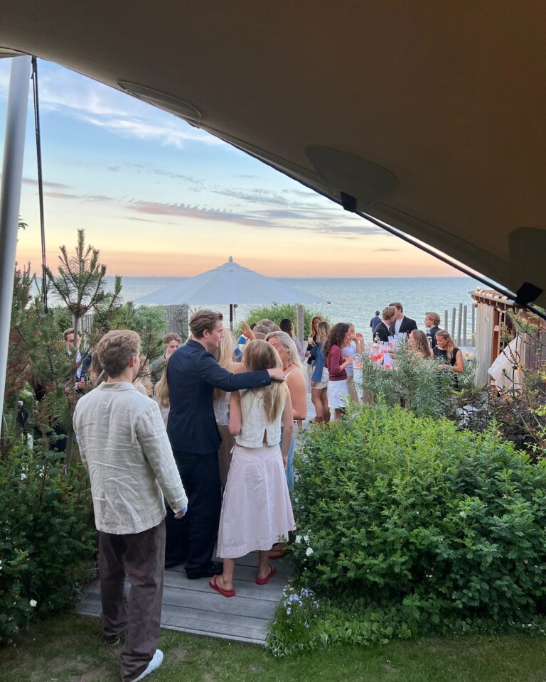 Guests socializing under tent at sunset waterfront event with scenic ocean backdrop, marquee structure, manicured landscape, and dramatic evening sky