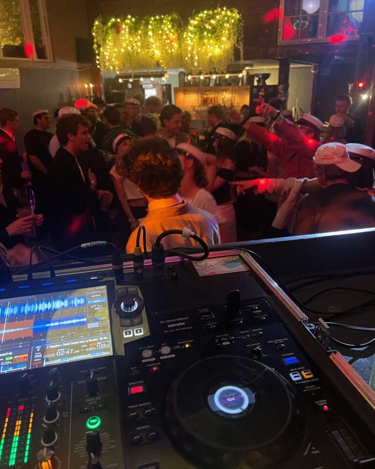 DJ mixer console at indoor venue with large dancing crowd, gold glitter backdrop overhead, red and warm lighting, packed dance floor with guests enjoying party