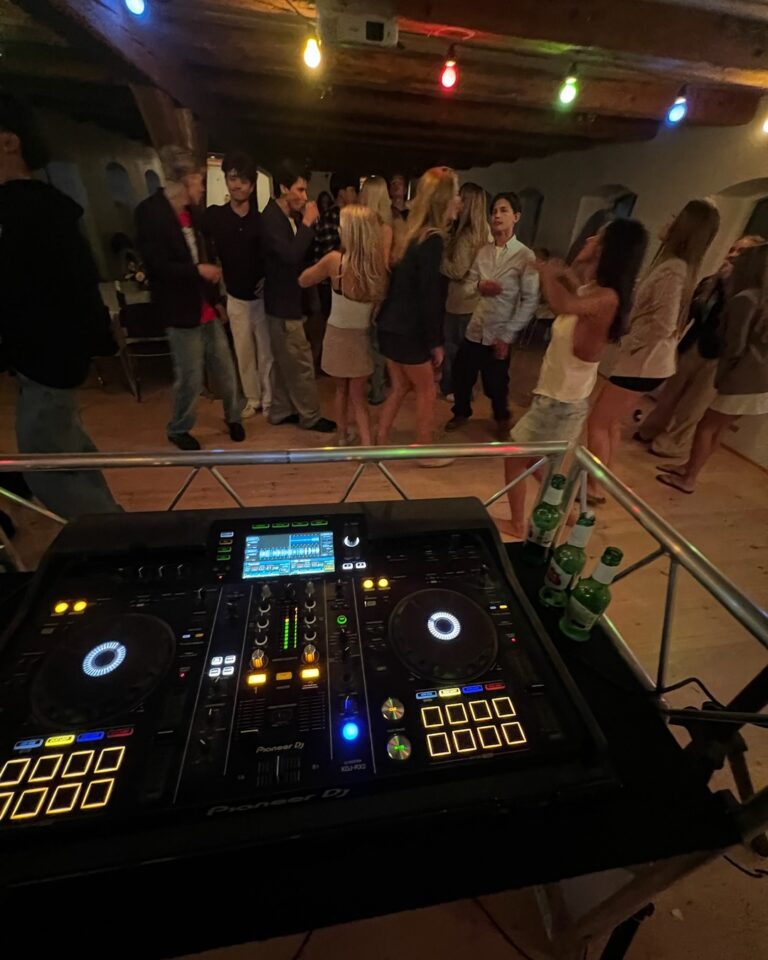 DJ mixer console at basement event venue with multicolor ceiling lighting, guests socializing and dancing on wooden floor with rustic exposed beam ceiling