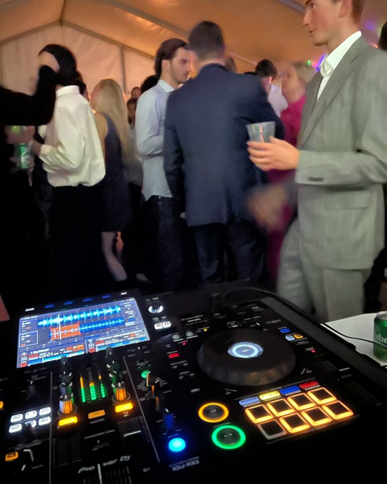 DJ mixing console with illuminated controls at formal event inside tent with well-dressed guests networking and socializing in business casual attire
