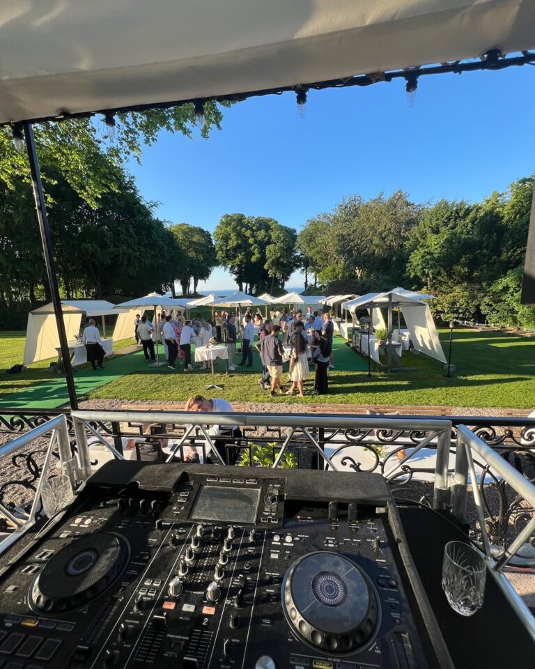 DJ mixer console at elevated platform overlooking luxury outdoor daytime event with multiple white tent pavilions, guests, manicured lawn, trees, and waterfront views